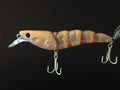 A close-up of a hard bait fishing lure with a realistic tan and brown striped body, a clear diving lip, and two silver treble hooks, set against a solid black background.