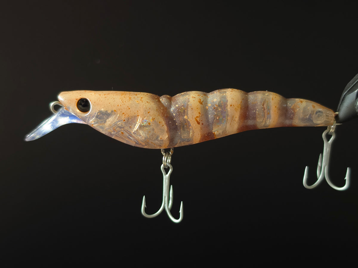 A close-up of a hard bait fishing lure with a realistic tan and brown striped body, a clear diving lip, and two silver treble hooks, set against a solid black background.