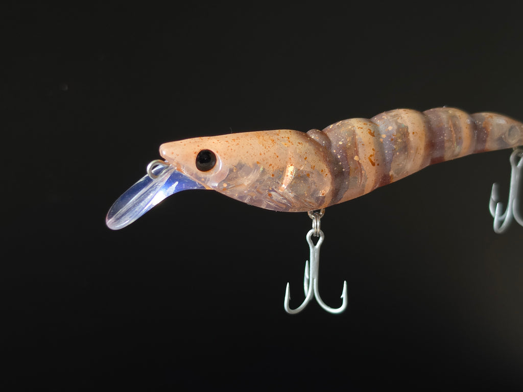 A close-up of a shrimp-shaped fishing lure with a clear diving lip and two metal treble hooks, shown against a solid black background.