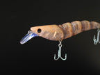 A close-up of a shrimp-shaped fishing lure with a clear diving lip and two metal treble hooks, shown against a solid black background.