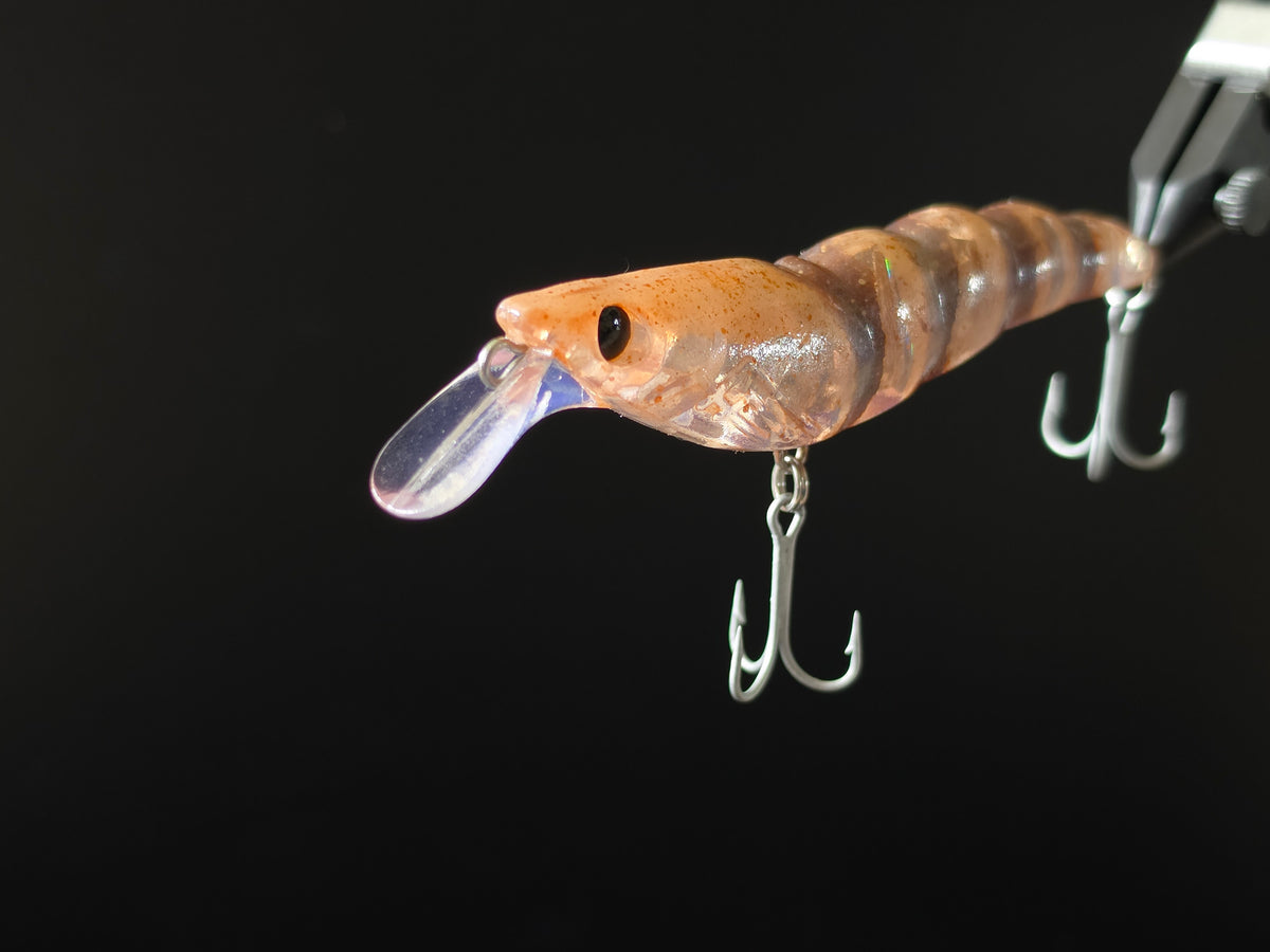 A close-up of a translucent brown shrimp-shaped fishing lure with a clear diving lip and silver treble hooks, held by forceps against a solid black background.