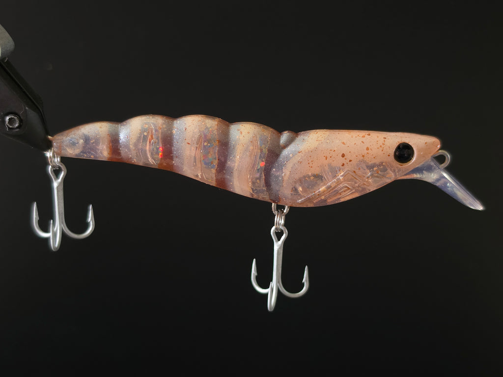 A close-up of a fishing lure with a translucent brown and beige body, subtle orange and blue iridescence, a clear plastic lip, and two silver treble hooks against a solid black background.