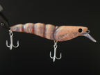 A close-up of a fishing lure with a translucent brown and beige body, subtle orange and blue iridescence, a clear plastic lip, and two silver treble hooks against a solid black background.
