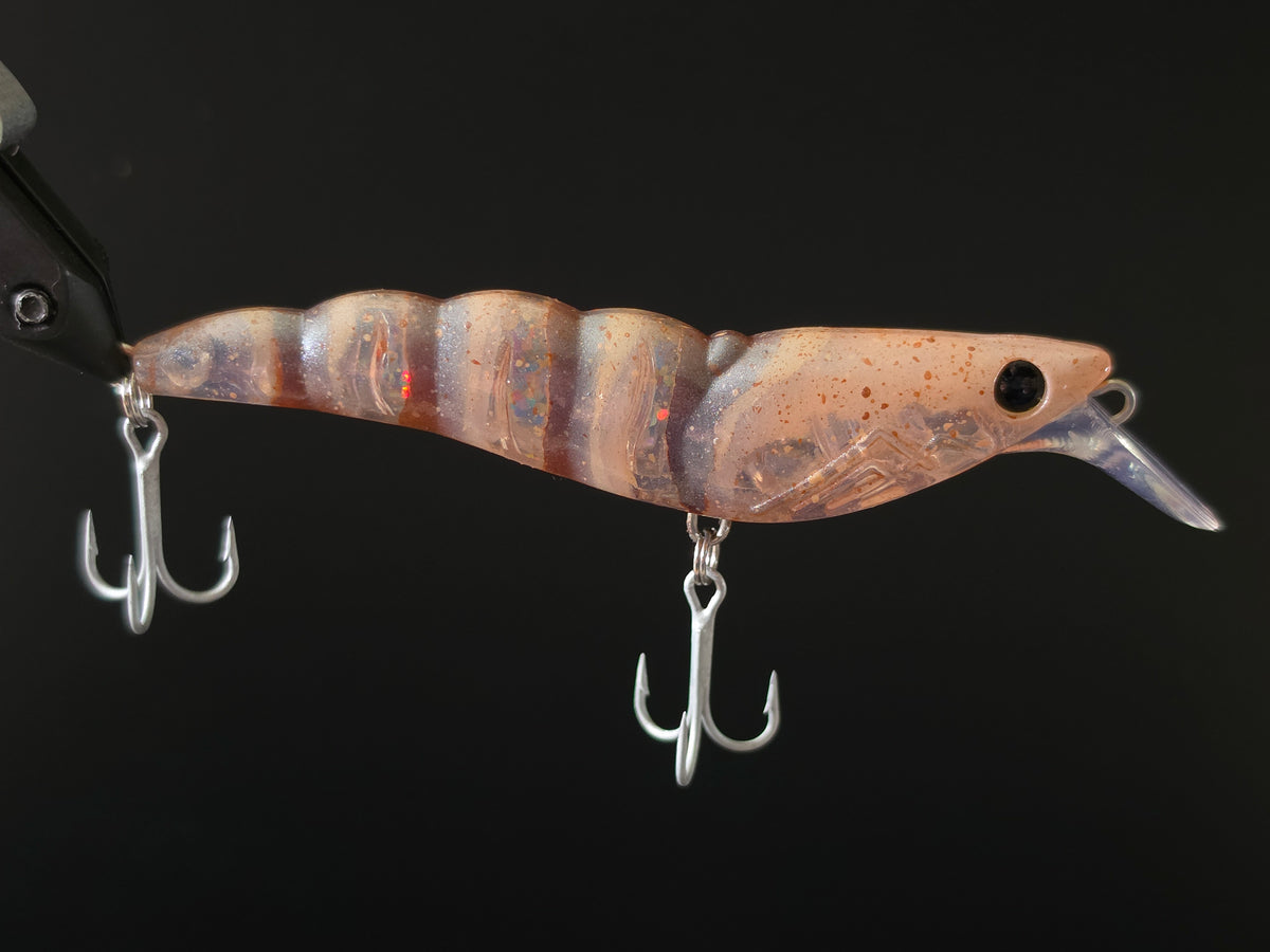 A close-up of a fishing lure with a translucent brown and beige body, subtle orange and blue iridescence, a clear plastic lip, and two silver treble hooks against a solid black background.