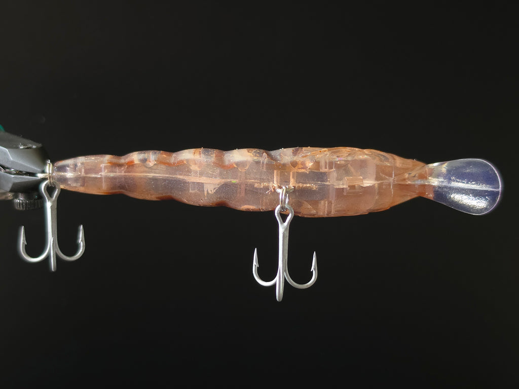 A close-up of a transparent swimbait fishing lure with a wavy body, orange-brown internal marbling, and two silver treble hooks shown against a solid black background.