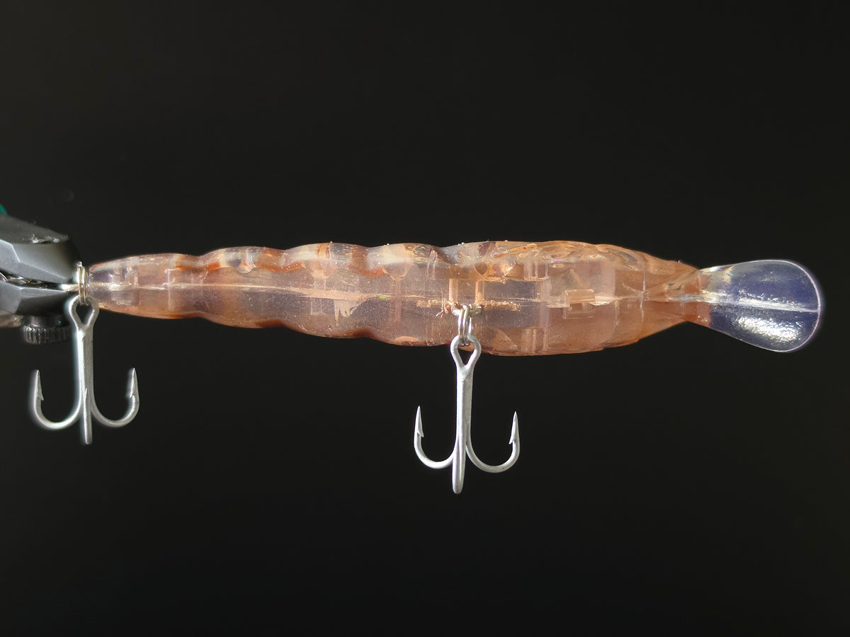 A close-up of a transparent swimbait fishing lure with a wavy body, orange-brown internal marbling, and two silver treble hooks shown against a solid black background.