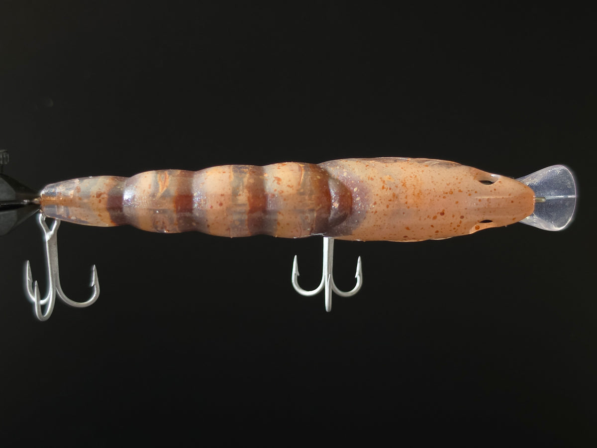 A hard bait fishing lure with a ribbed, shrimp-like body in brown and beige tones, featuring a clear plastic tail and two silver treble hooks, shown against a solid black background.