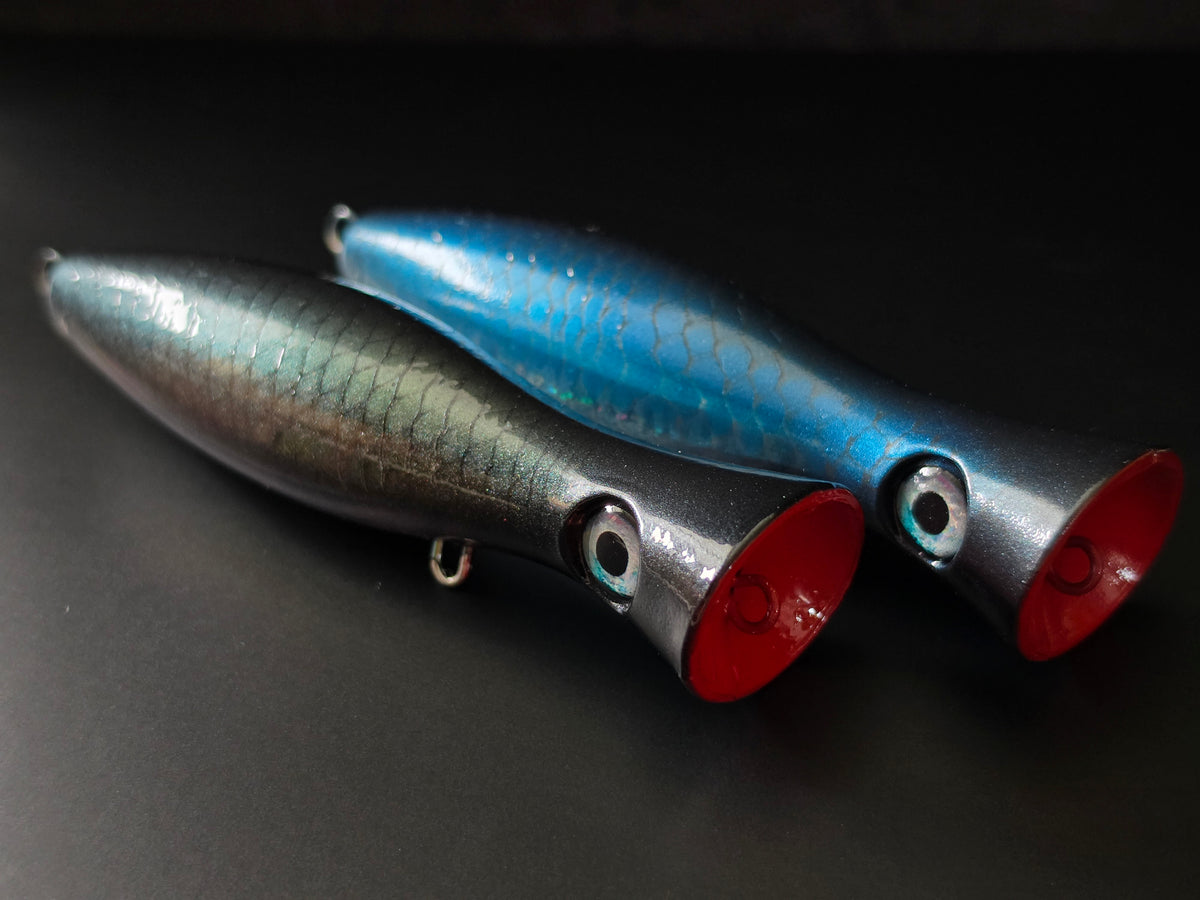 Two fishing lures on a dark background, one with a metallic black and green scale pattern and red mouth, and the other with a metallic blue and black scale pattern and red mouth.