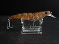 An orange-brown translucent shrimp fishing lure with a clear diving lip and silver treble hooks, displayed on a clear acrylic stand against a dark background.