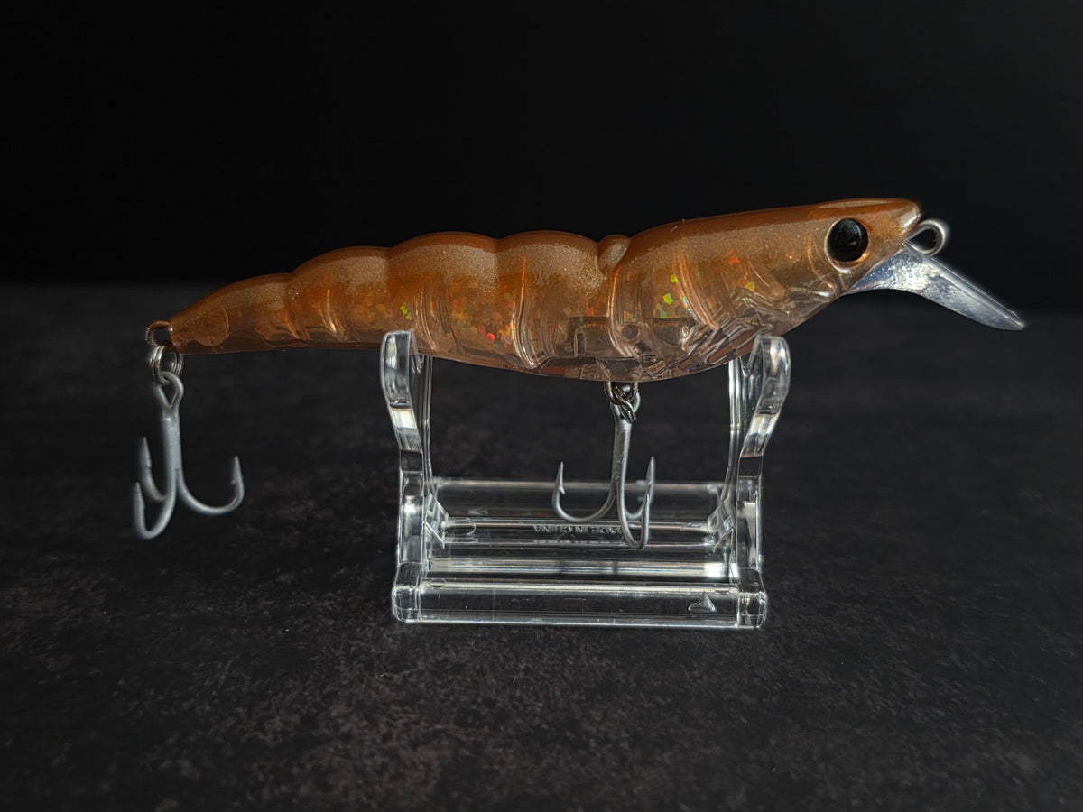 An orange-brown translucent shrimp fishing lure with a clear diving lip and silver treble hooks, displayed on a clear acrylic stand against a dark background.