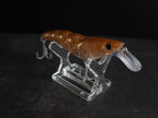 A brown translucent soft plastic fishing lure with a clear diving lip and two treble hooks, displayed on a clear acrylic stand against a dark background.