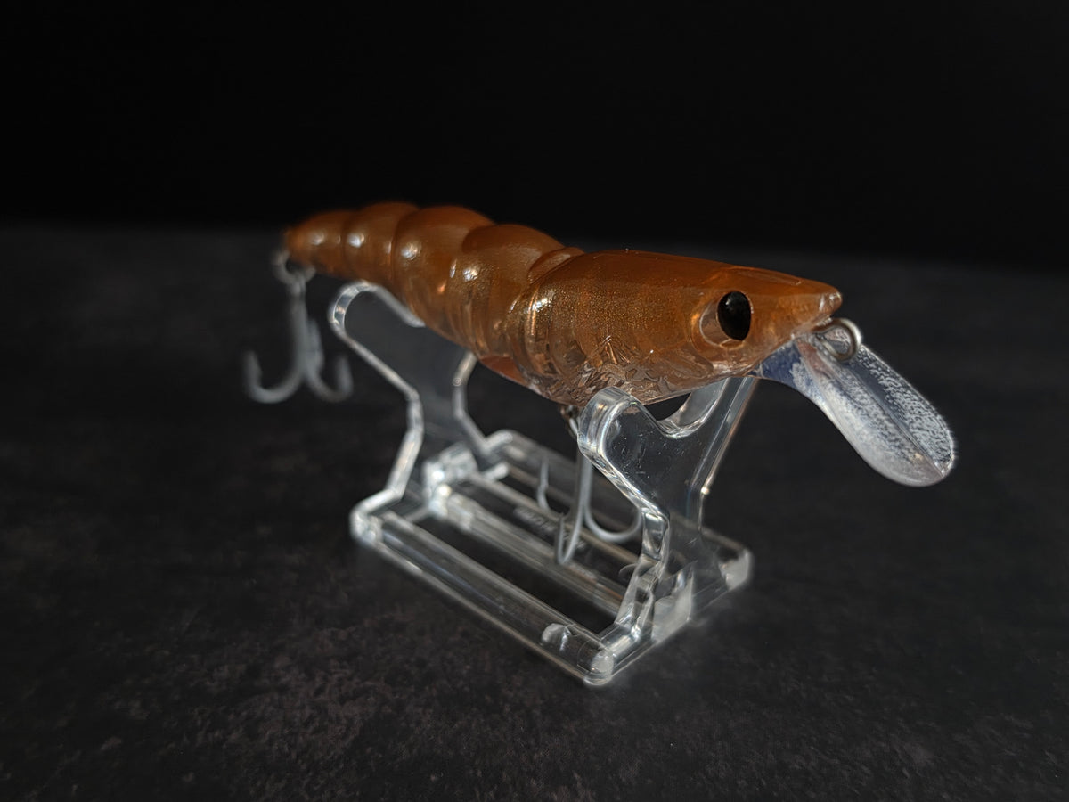 A brown translucent soft plastic fishing lure with a clear diving lip and two treble hooks, displayed on a clear acrylic stand against a dark background.