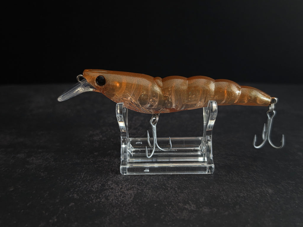An amber-brown translucent hard bait fishing lure with a clear diving lip and silver treble hooks, displayed on a clear acrylic stand against a dark background.