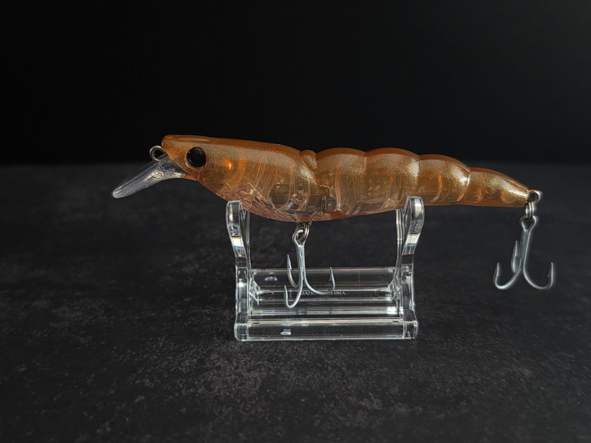 An amber-brown translucent hard bait fishing lure with a clear diving lip and silver treble hooks, displayed on a clear acrylic stand against a dark background.