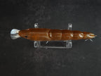 A brown translucent hard plastic fishing lure with silver metal hooks displayed horizontally on a clear acrylic stand against a dark textured background.