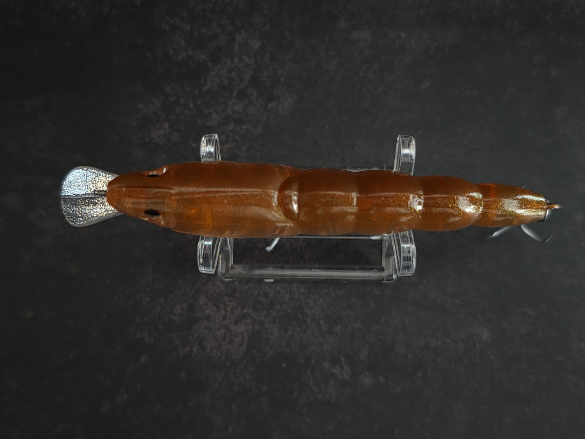 A brown translucent hard plastic fishing lure with silver metal hooks displayed horizontally on a clear acrylic stand against a dark textured background.