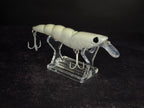 A white plastic fishing shrimp lure with a clear plastic diving lip and two treble hooks, displayed on a clear acrylic stand against a dark background.