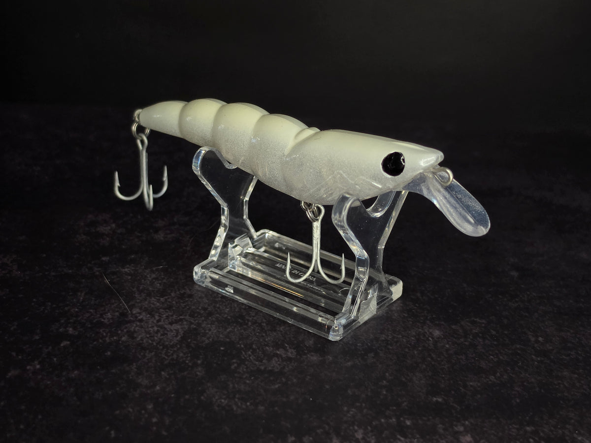 A white plastic fishing shrimp lure with a clear plastic diving lip and two treble hooks, displayed on a clear acrylic stand against a dark background.