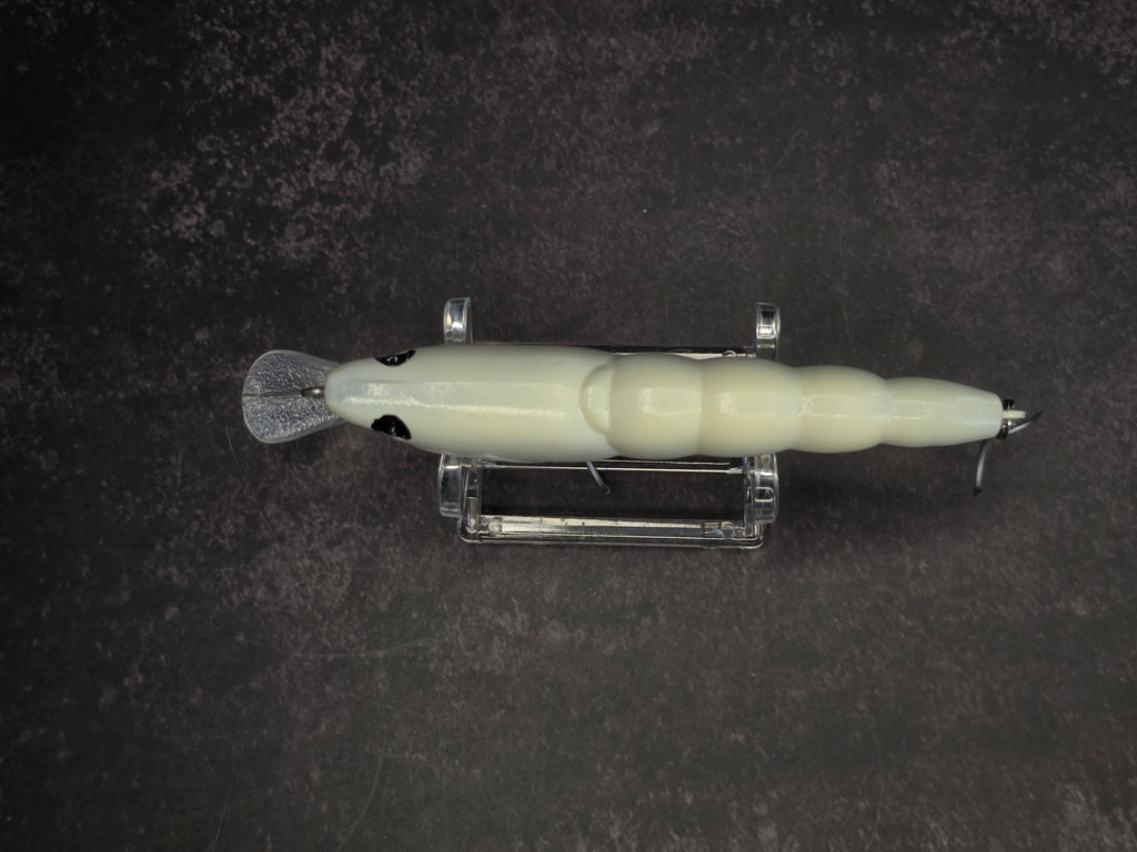 A pale cream-colored fishing lure with a clear plastic tail and metal hooks, displayed horizontally on a clear stand against a dark textured background.