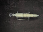 A pale cream-colored fishing lure with a clear plastic tail and metal hooks, displayed horizontally on a clear stand against a dark textured background.