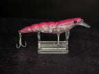 A pink and iridescent fishing lure with two treble hooks, displayed on a clear acrylic stand against a black background.