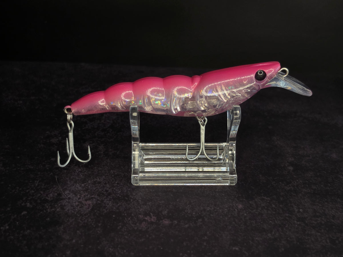A pink and iridescent fishing lure with two treble hooks, displayed on a clear acrylic stand against a black background.