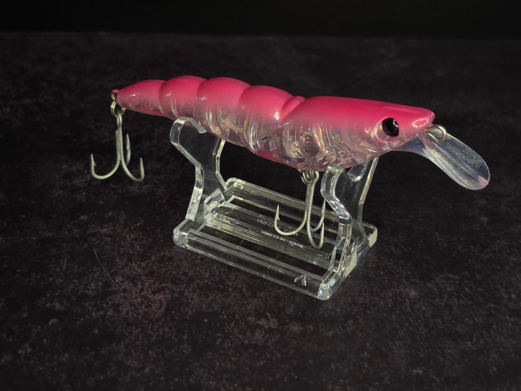 A pink and clear plastic fishing lure with a clear diving lip and metal treble hooks, displayed on a clear acrylic stand against a dark textured background.