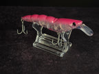A pink and clear plastic fishing lure with a clear diving lip and metal treble hooks, displayed on a clear acrylic stand against a dark textured background.