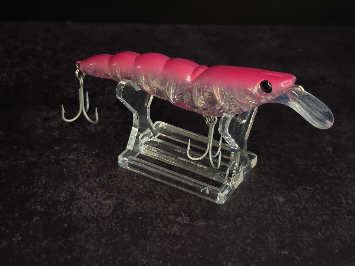 A pink and clear plastic fishing lure with a clear diving lip and metal treble hooks, displayed on a clear acrylic stand against a dark textured background.