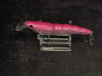 A pink hard bait fishing lure with a clear diving lip and metal treble hooks, displayed on a clear acrylic stand against a dark textured background.