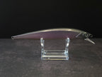 A metallic black and purple fishing lure with a realistic body, displayed on a clear acrylic stand against a dark background.
