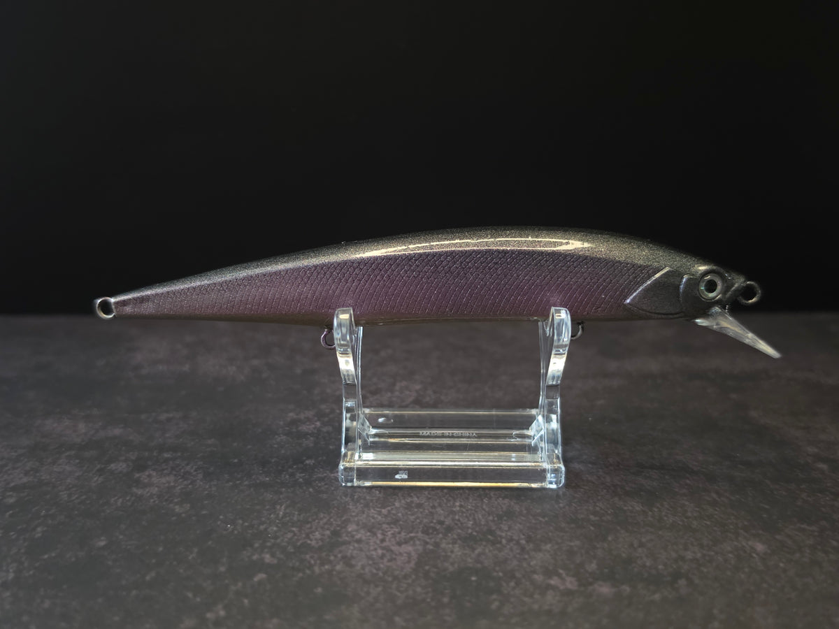 A metallic black and purple fishing lure with a realistic body, displayed on a clear acrylic stand against a dark background.
