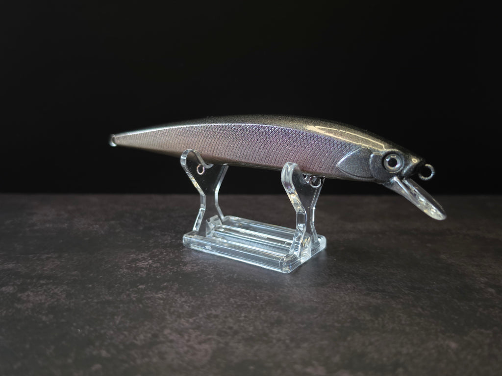 A black and purple hard bait fishing lure with a realistic fish body, standing on a clear acrylic display stand against a dark background.