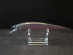 A metallic purple and black fishing lure with a realistic fish body, displayed on a clear acrylic stand against a dark background.