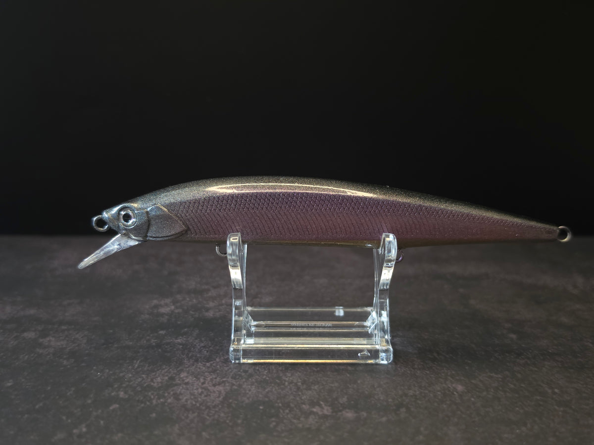 A metallic purple and black fishing lure with a realistic fish body, displayed on a clear acrylic stand against a dark background.