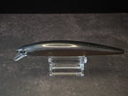 A dark metallic black hard bait fishing lure with a clear plastic diving lip, displayed on a clear acrylic stand against a dark textured background.