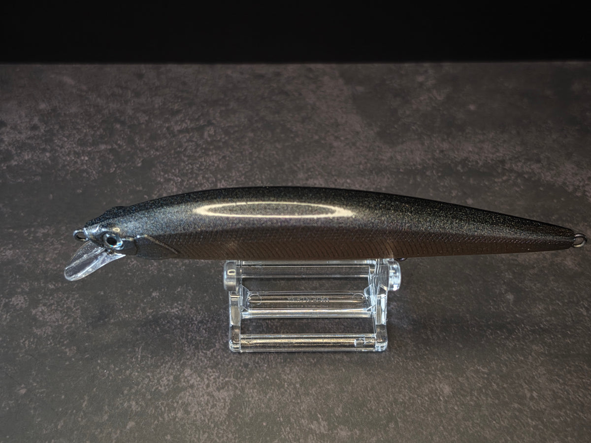 A dark metallic black hard bait fishing lure with a clear plastic diving lip, displayed on a clear acrylic stand against a dark textured background.
