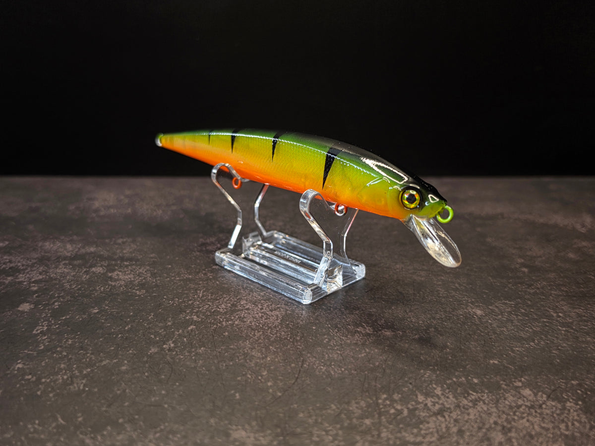 A hard bait fishing lure with a yellow-to-orange gradient body, black tiger-like stripes, and a clear diving lip, displayed on a clear acrylic stand against a dark background.