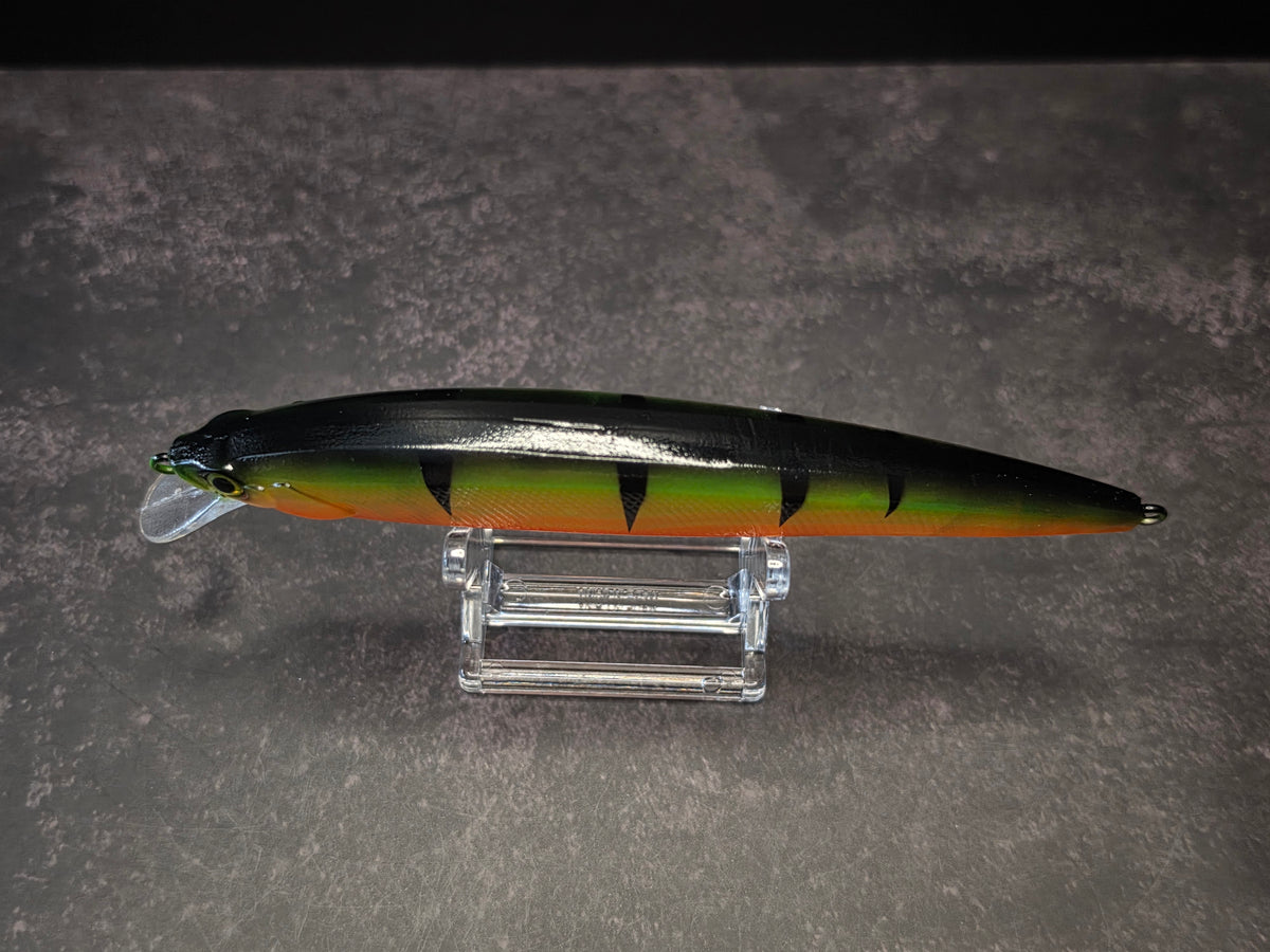 A hard plastic fishing lure with a black, green, and orange tiger stripe pattern, displayed on a clear acrylic stand against a textured gray background.