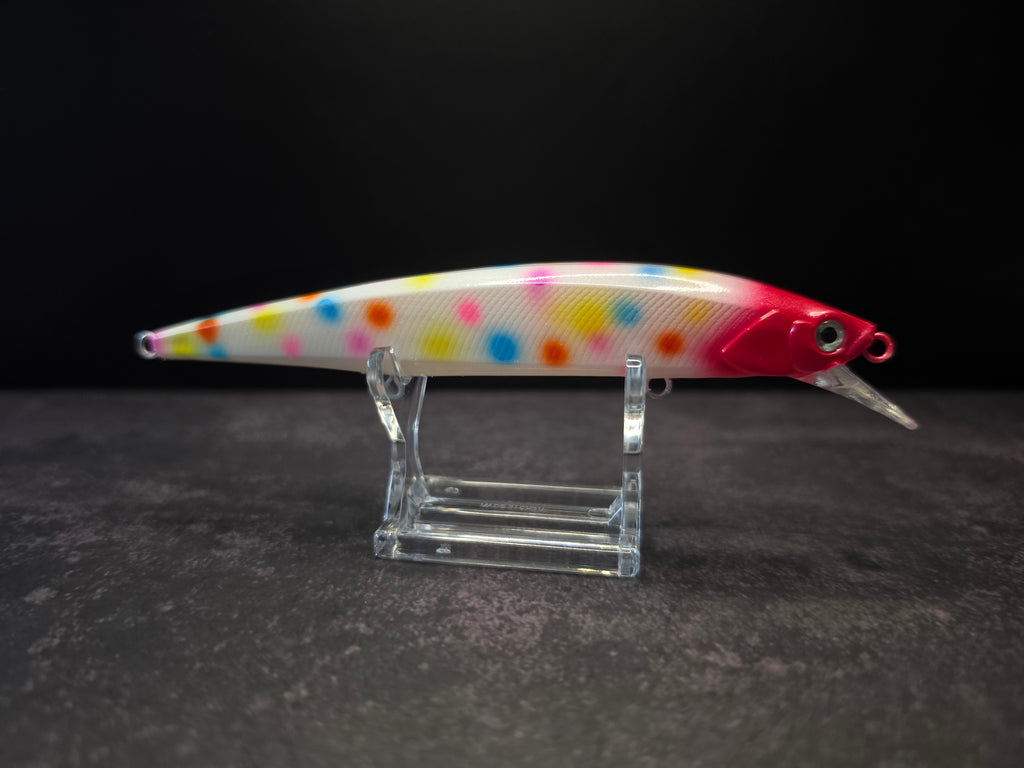 A hard bait fishing lure with a white body covered in colorful polka dots, featuring a bright red head and a clear plastic diving lip, displayed on a clear acrylic stand against a dark background.
