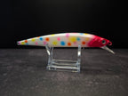A hard bait fishing lure with a white body covered in colorful polka dots, featuring a bright red head and a clear plastic diving lip, displayed on a clear acrylic stand against a dark background.