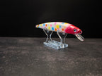 A hard bait fishing lure with a white body covered in colorful polka dots and a red head, displayed on a clear acrylic stand against a dark background.
