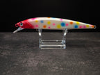 A hard bait fishing lure with a white body, red head, and multicolor polka dots, displayed on a clear acrylic stand against a dark background.