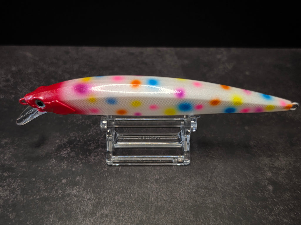 A white fishing lure with colorful polka dots and a red head, displayed on a clear acrylic stand against a dark gray background.