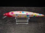 A white fishing lure with colorful polka dots and a red head, displayed on a clear acrylic stand against a dark gray background.