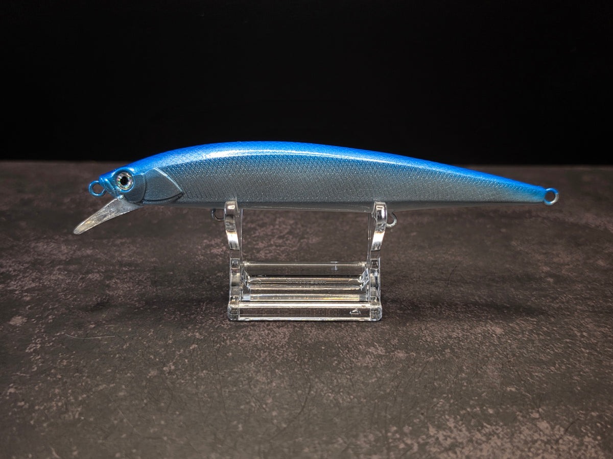 A blue and silver hard bait fishing lure with a clear plastic diving lip, displayed on a clear acrylic stand against a dark background.