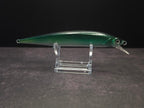 A green metallic fishing lure with a clear plastic diving lip, displayed on a clear acrylic stand against a dark background.