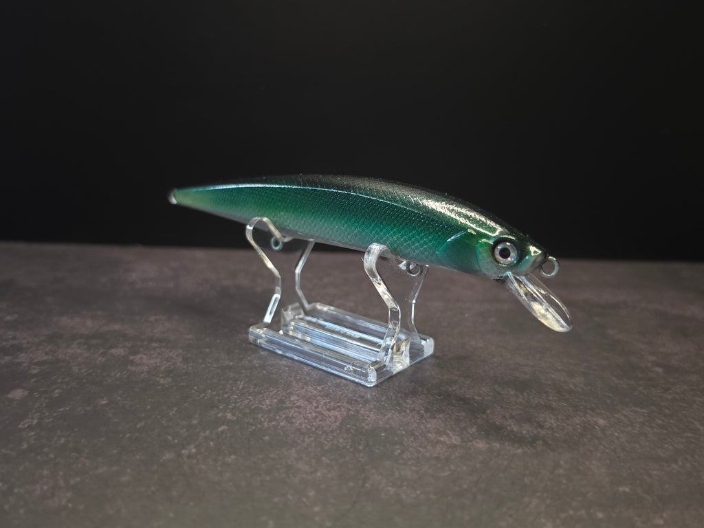 A metallic green and black hard bait fishing lure with a clear plastic lip, displayed on a clear acrylic stand against a dark background.