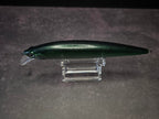 A metallic dark green fishing lure with a clear diving lip, displayed on a clear acrylic stand against a textured gray background.