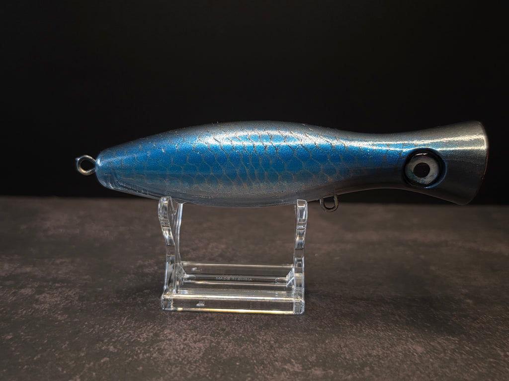 A metallic black and blue fishing lure with a red mouth, displayed on a clear acrylic stand against a dark background.